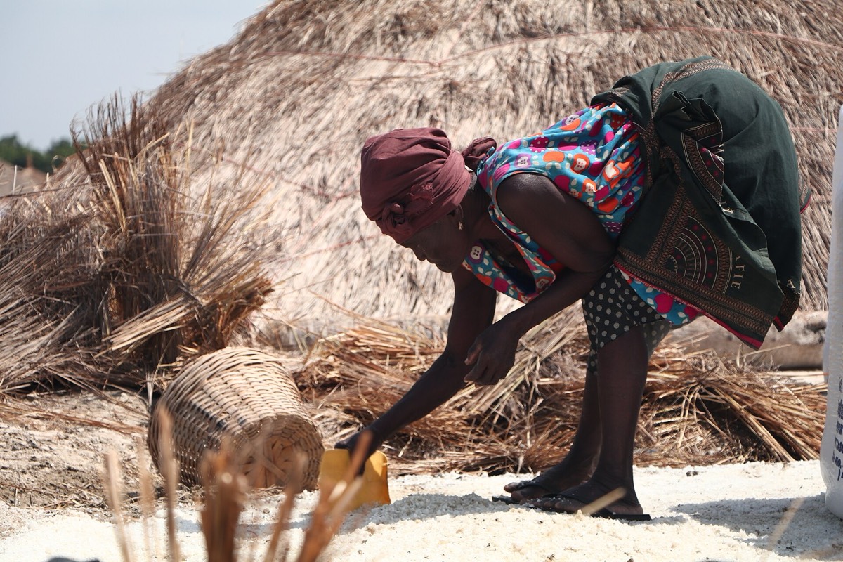 A woman in a headwrap bends to gather salt beside a thatched salt mound on the Songor Lagoon